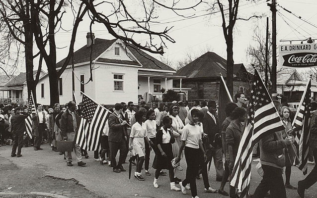 B&W; marchers with American flags walk in front of a white house. Coca-cola sign hangs on right. B&W; marchers with American flags walk in front of a white house. Coca-cola sign hangs on right.