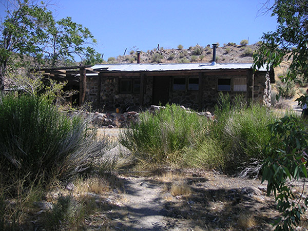 A single-story rustic cabin is constructed of stone and wood, surrounded by a dry landscape.