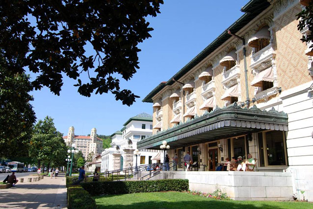 People sit under an awning at the front of an ornate brick building, with hedgerow in front.