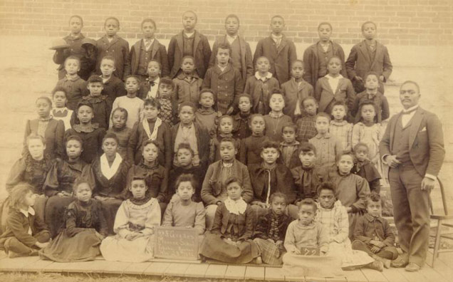 A sepia-toned class portrait with six rows of children and a male teacher in a suit. A sepia-toned class portrait with six rows of children and a male teacher in a suit.