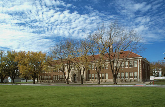 A long, two-story brick school is surrounded by lawn, a row of trees, and blue sky. 