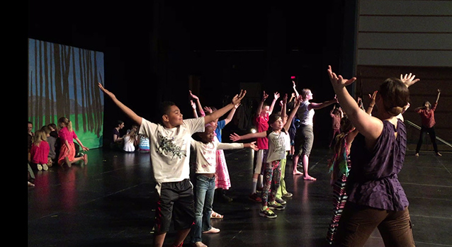Students stand on stage raising their arms high above their heads. Students stand on stage raising their arms high above their heads.