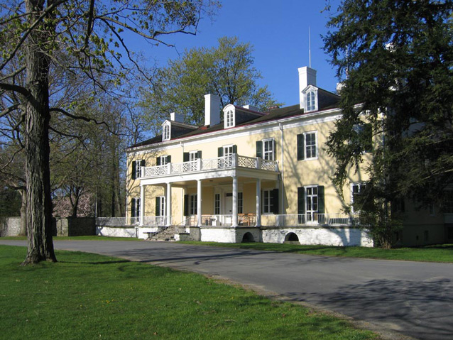 A driveway curves in front of a sprawling, two story yellow house with many windows.