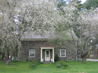 Trees bloom outside a two-story field stone structure, with a door framed by two windows.