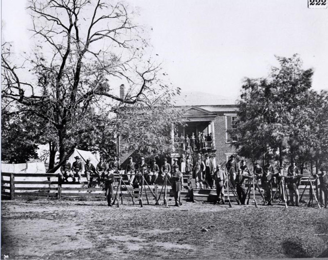 Soldiers pose with their guns along the wooden fence in front of the brick Court House. 