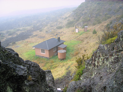 A high-angle view from a rocky cliff towards a simple, one-story cabin and water tank.