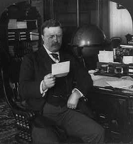 Black and white photo of President Theodore Roosevelt sitting at his desk.  