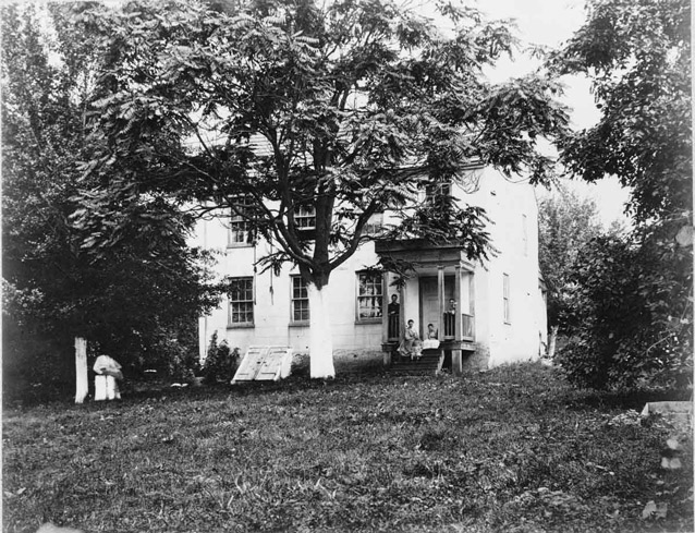 People sit on the front porch of a rectangular white farmhouse, surrounded by trees and yard.