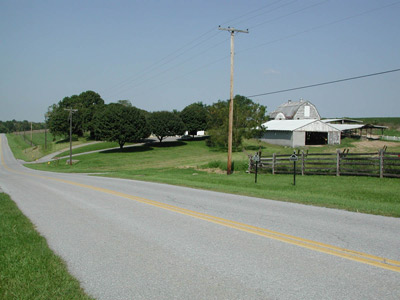 A straight road passes over gently rolling terrain, past a cluster of farm structures and trees.