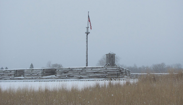 A light snow dusts the logs of a fort, with an American flag that thrusts up into the gray sky. 