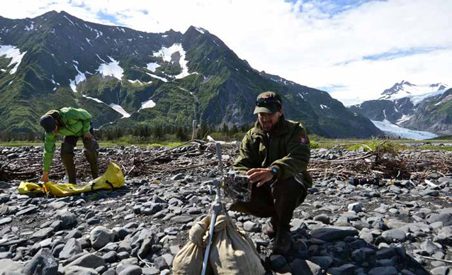 two men, one in a ranger uniform, set up a camera on a rocky beach