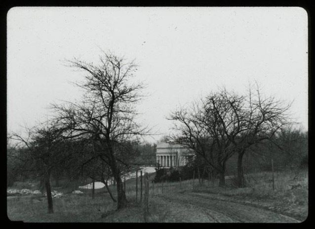 The white columns of the memorial stand out in the distance, beyond bare trees and a dirt road.