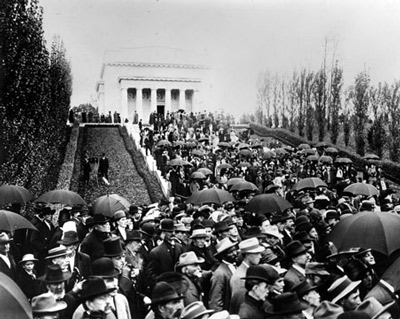 A crowd of people, many with umbrellas and hats, gather on the stairs to a neoclassical building.