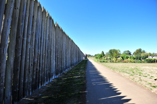 A dirt path follows the right edge of a stockade wall, half in shadow from the tall timbers. 