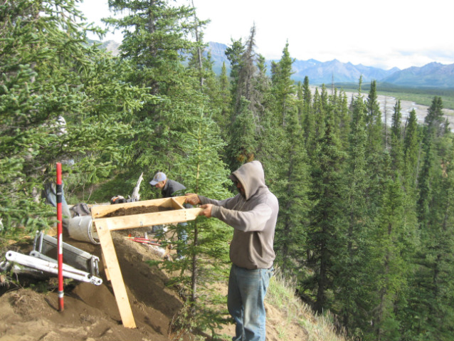 man sifts through dirt on a hill with mountains and spruce trees in the background man sifts through dirt on a hill with mountains and spruce trees in the background
