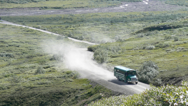 a green bus drives down a gravel road with dust filling the air behind it