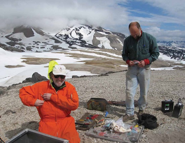 two men stand on a gravelly surface with snowy mountains in the distance two men stand on a gravelly surface with snowy mountains in the distance