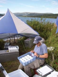 researcher sits surrounded by field equipment next to a lake