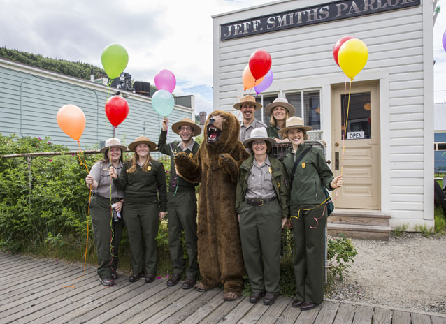 Park rangers and person in bear costume hold balloons in front of building. 