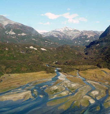 view of braided river with shrub-covered hills and rocky mountains in the distance
