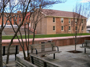 A area with trees and park benches with a two-story brick building in the background