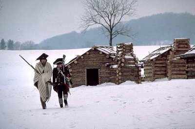 Two Revolutionary War reenactors walk in the snow with a log cabin and trees behind them Two Revolutionary War reenactors walk in the snow with a log cabin and trees behind them