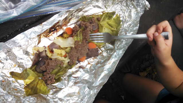 A child uses a fork to scoop a bite of cooked carrots, beef, potatoes, and cabbage on aluminum foil. A child uses a fork to scoop a bite of cooked carrots, beef, potatoes, and cabbage on aluminum foil.