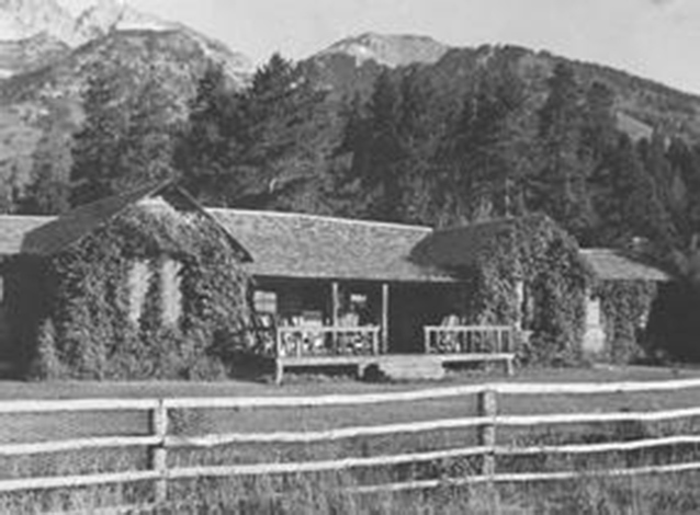 Log cabin at the base of mountains, with vines growing on the front and a wooden fence in front