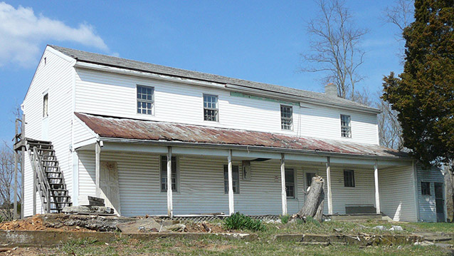 A rectangular, two-story house with wide siding, small windows, and an awning across the front.