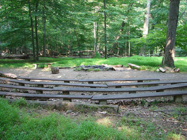 Wooden benches form a semi-circle around a campfire area, surrounded by forest Wooden benches form a semi-circle around a campfire area, surrounded by forest