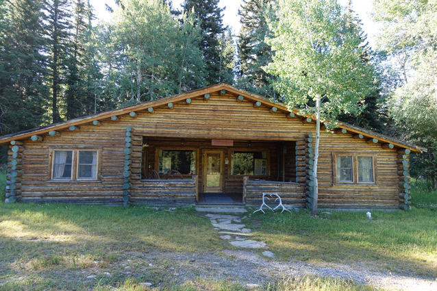 A square log cabin has a sloping roof and sheltered front porch.