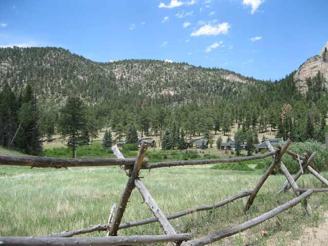 A wooden fence frames the landscape, including grassy area, ranch buildings, and pine-covered hills.