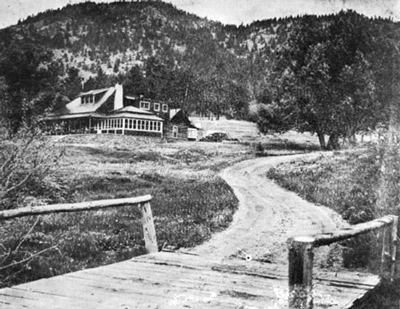 Historic photo of a narrow wooden bridge and dirt road leading to a large ranch house.