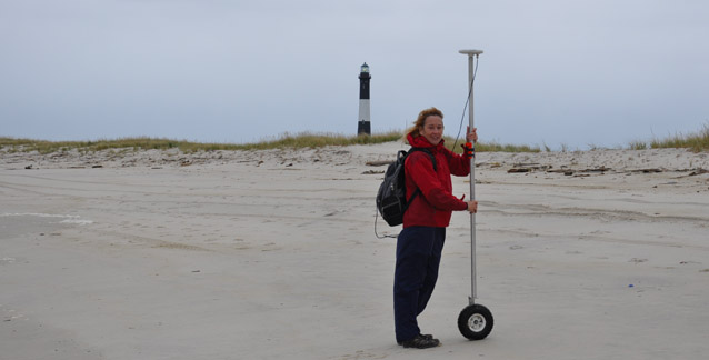 A scientist holds a GPS unit on a pole with a wheel on the beach.