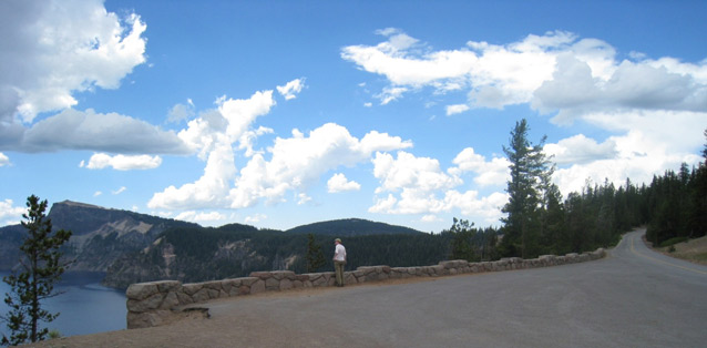 A person stands beside a low stone guardwall, overlooking a dramatic view of a lake and mountains.