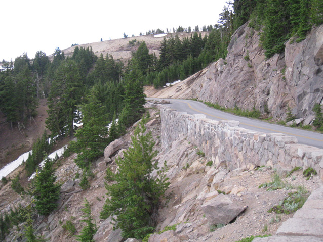 Subalpine forest grows along a rocky slope, where a stone wall holds up a road against the hillside.