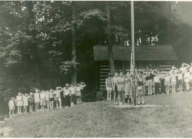 A crowd of young people, near a cabin, salutes as several girls attach the flag to a pole. A crowd of young people, near a cabin, salutes as several girls attach the flag to a pole.