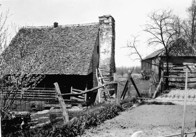 Historic photo of a log farm house with steep, shingled roof, stone chimney, and wooden fence.
