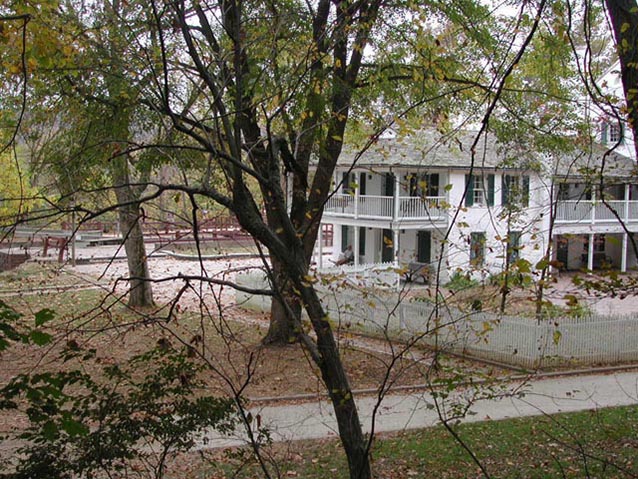 The two-story Tavern with porches and picket fence, seen through trees. The two-story Tavern with porches and picket fence, seen through trees.