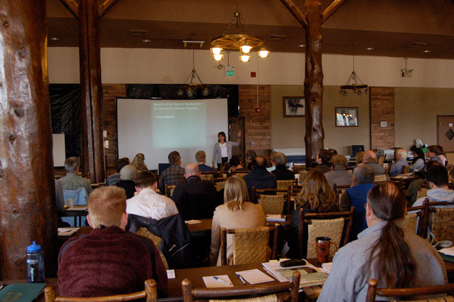 A woman stands in front of a projection screen and addresses a seated crowd in a large, rustic room.