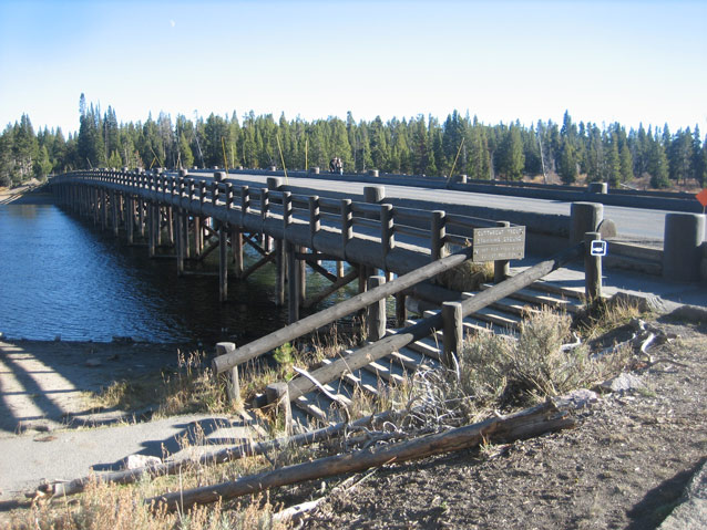 A thick wooden railing lines the side of a long bridge over a blue river.