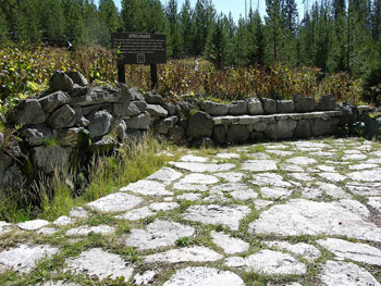 A patio paved with large stones in front of a stone bench and stacked stone wall, framed by conifers