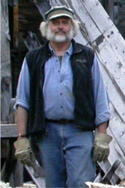 A man with a grey beard, work gloves, cap, blue jeans, and unzipped fleece vest.