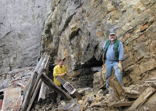 Two men stand beside a rough rock face, with wood and metal debris scattered around.