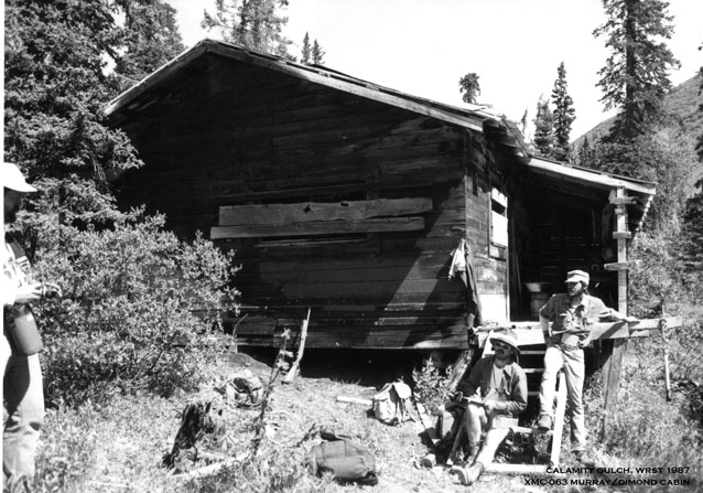 Three men with hats on the front steps of a wood cabin, surrounded by trees and hills