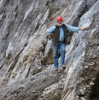A man in a red helmet stands with one hand on his hip and the other against a rock face