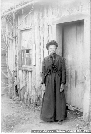 Woman wearing a heavy, long black dress and jacket and hat stands next to a log structure.