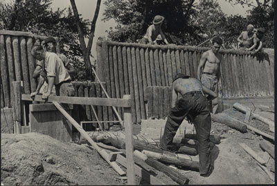 A crew of young men in work attire with scattered logs and a partially-built log fortification