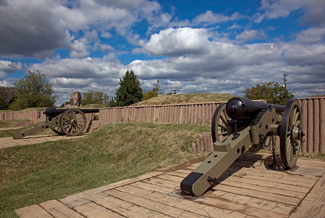 Puffy white clouds hang over two cannons, wooden fortifications, and earthworks.