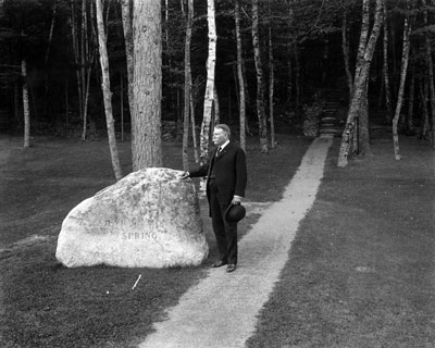 A man in a dark suit, holding a bowler hat, stands with one hand on a large boulder. A man in a dark suit, holding a bowler hat, stands with one hand on a large boulder.